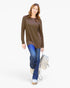A woman modeling a brown long-sleeve tunic top with a chest pocket, paired with blue jeans, beige sneakers, and carrying a light beige backpack against a plain white studio background.
