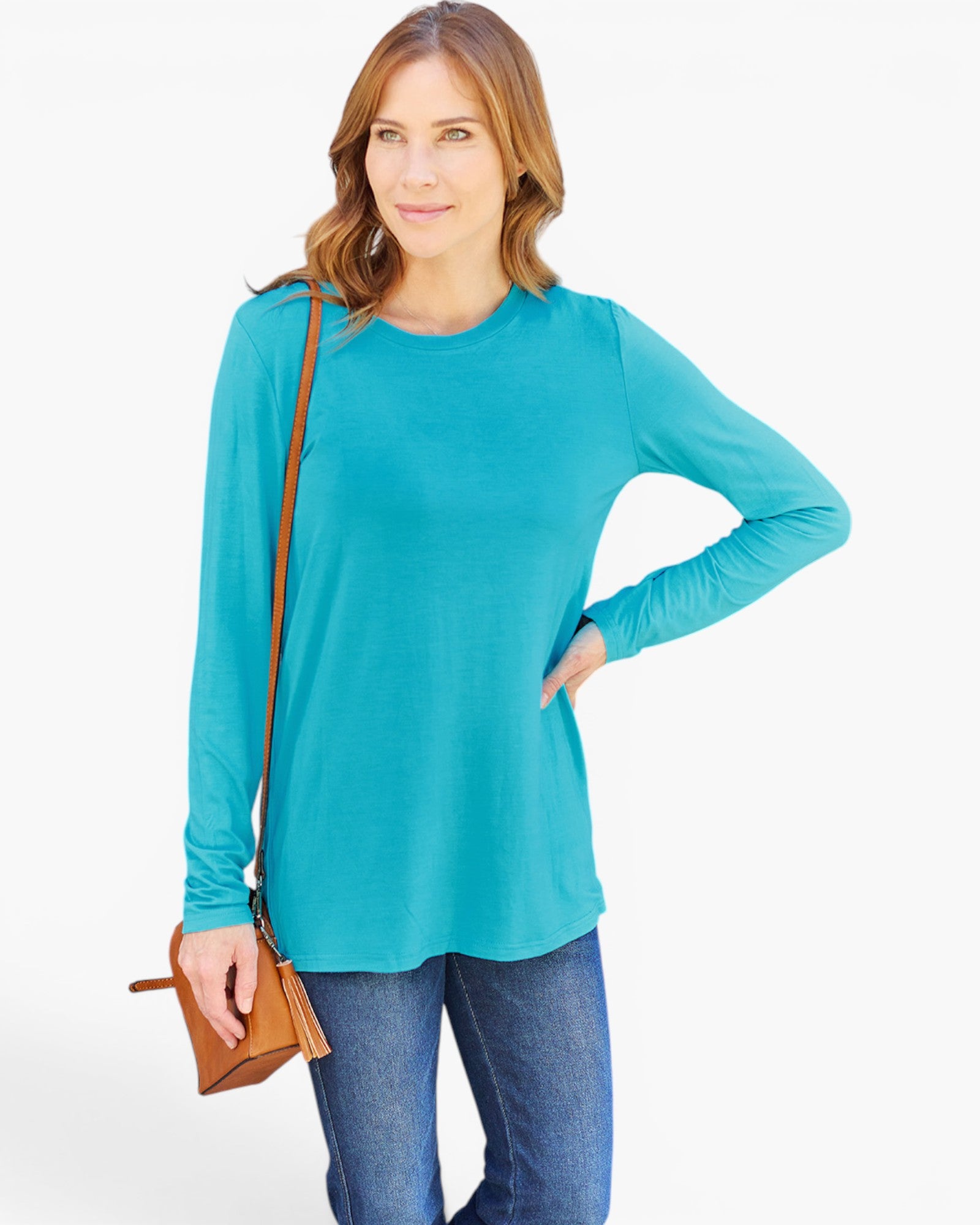 A woman modeling a turquoise long-sleeve crewneck top with blue jeans and a brown crossbody bag, set against a plain white studio background.