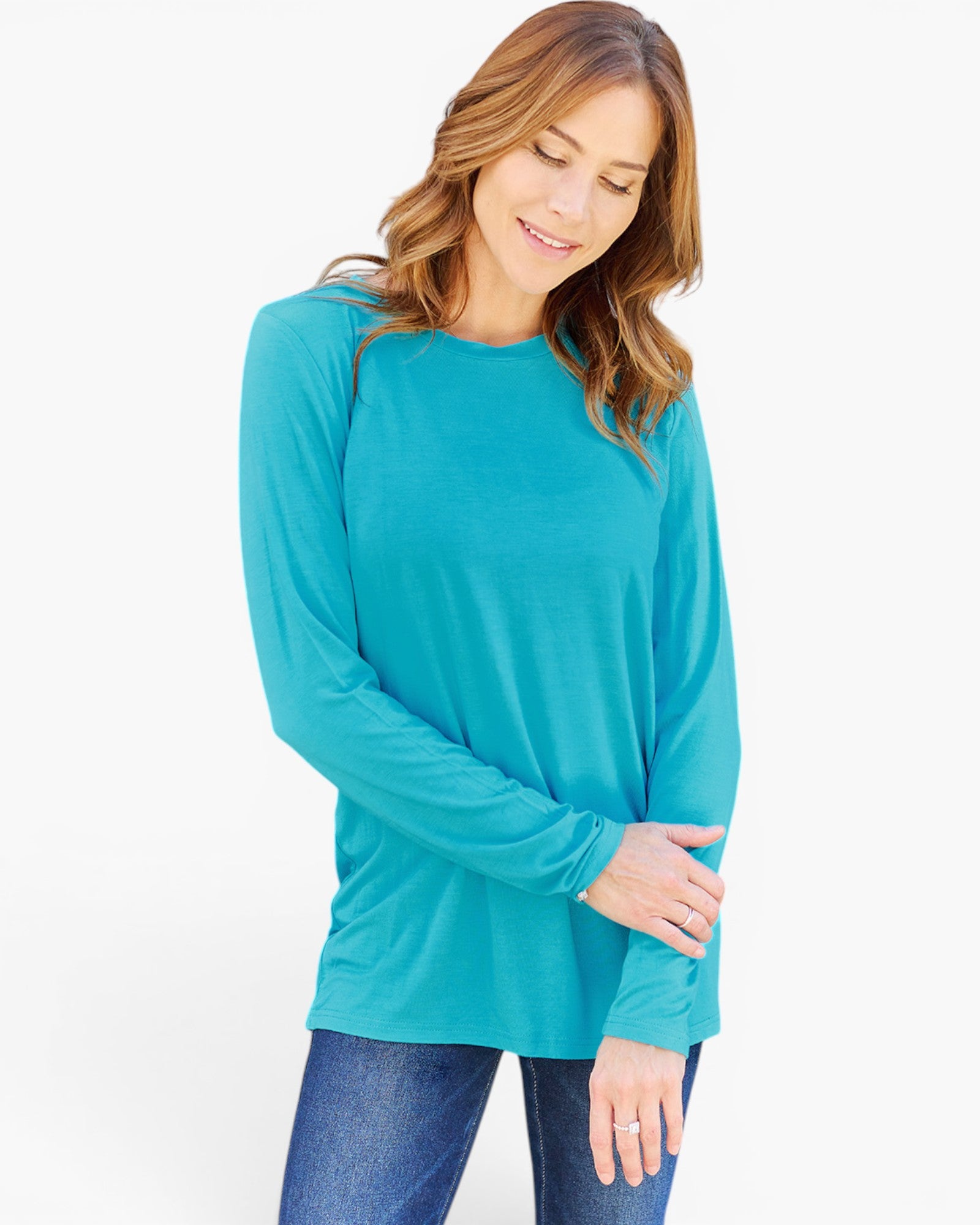 A woman wearing a bright turquoise long-sleeve crewneck top and blue jeans, standing against a plain white studio background.