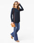 A woman modeling a black long-sleeve top with blue flared jeans, wearing sunglasses and carrying a small tan crossbody bag, set against a plain white studio background.