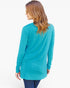 A woman with long wavy light brown hair is shown from the back wearing a bright turquoise long-sleeve tunic top and dark blue jeans against a plain white background.