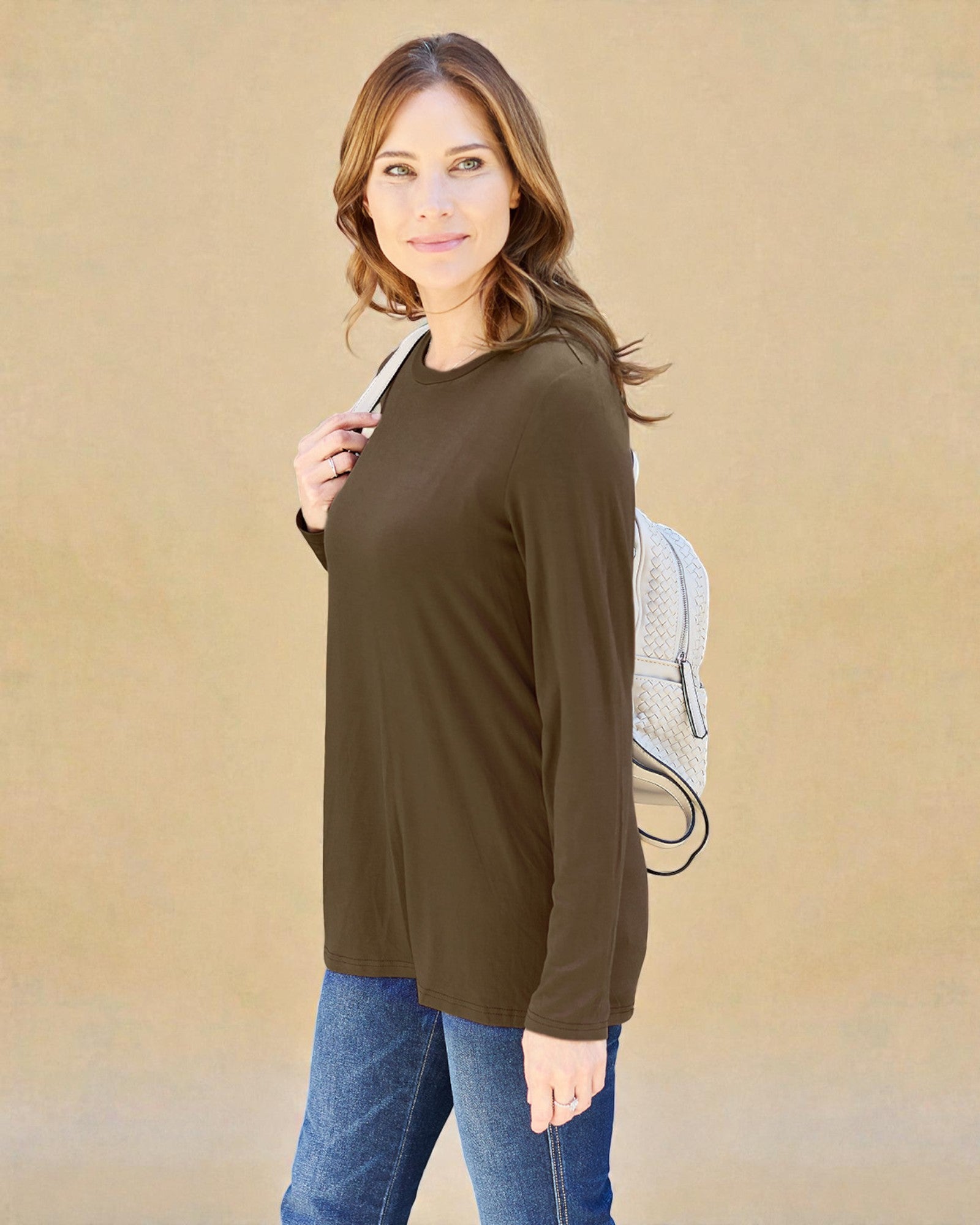 Woman wearing an olive green long-sleeve top and blue jeans against a beige background