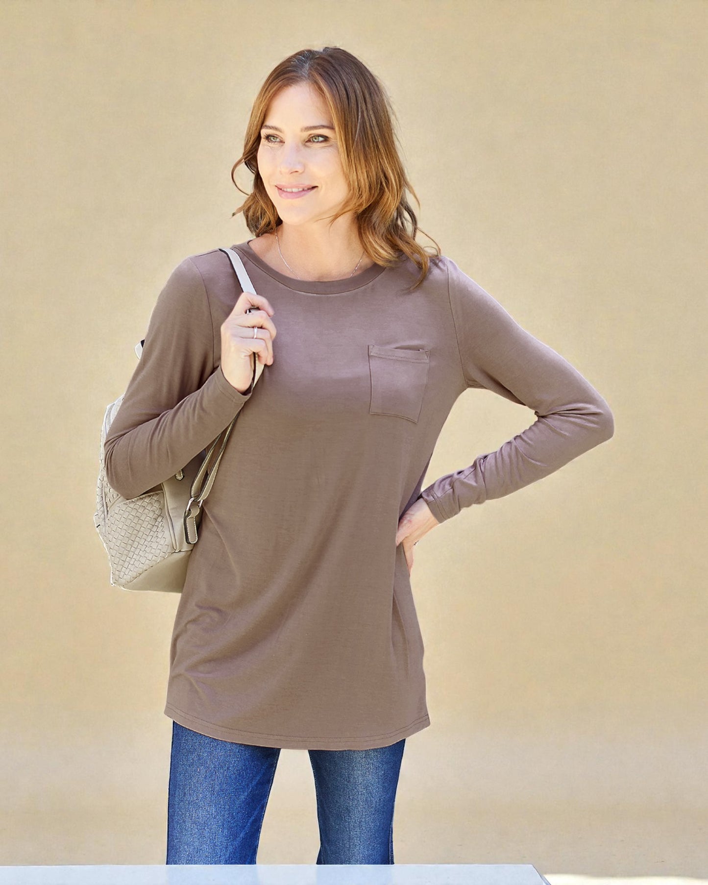 Woman wearing a long-sleeve brown top with a pocket, holding a beige bag against a beige background
