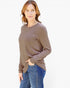 A woman modeling a long-sleeve crewneck top in taupe brown, paired with blue jeans, against a plain white background.