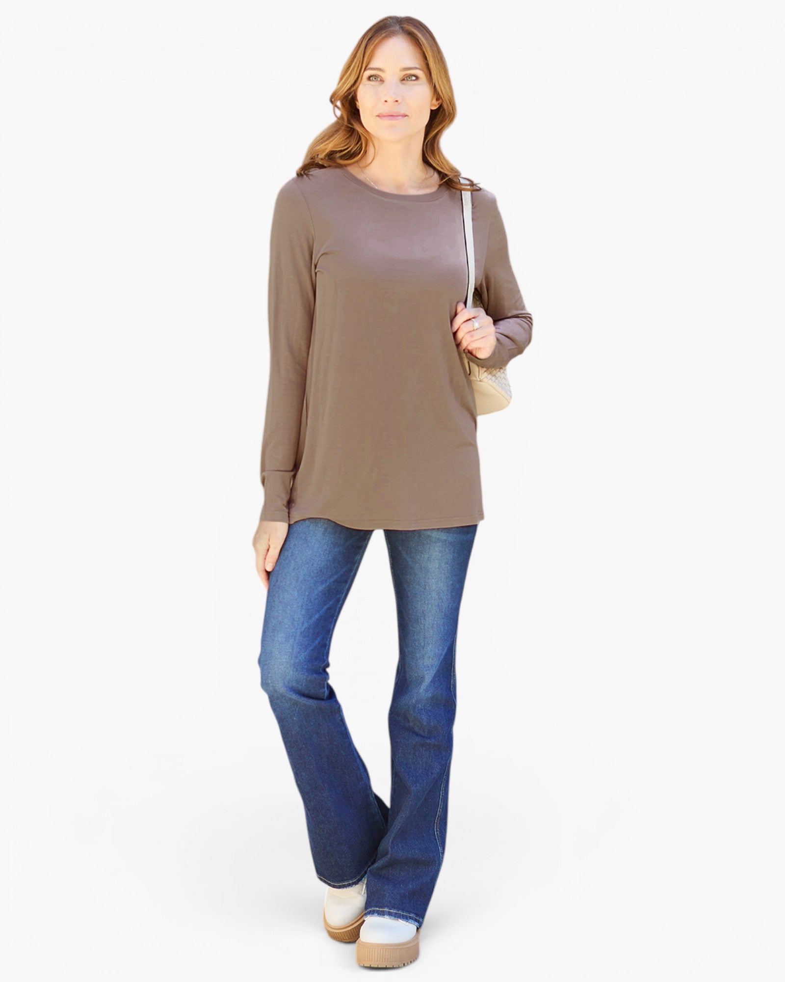 A woman modeling a taupe brown long-sleeve tunic top with blue jeans and a beige shoulder bag, standing against a plain white studio background.