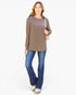 A woman modeling a taupe brown long-sleeve tunic top with blue jeans and a beige shoulder bag, standing against a plain white studio background.