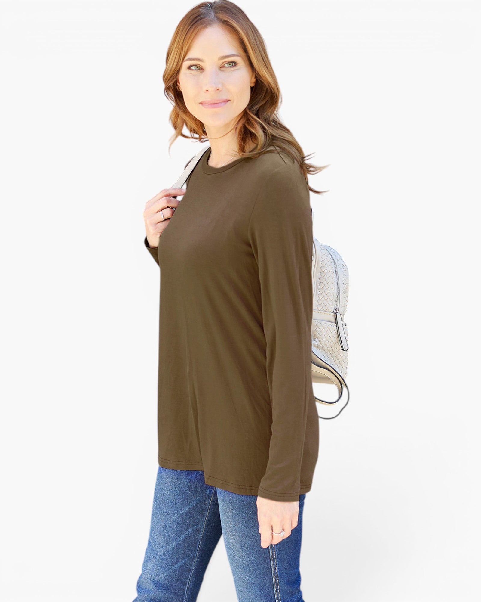 A woman modeling a long-sleeve crewneck top in olive brown, paired with blue jeans and a light-colored backpack, against a plain white studio background.