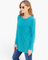 A woman modeling a teal long-sleeve tunic top with a single chest pocket, paired with blue jeans and round sunglasses, against a plain white studio background.