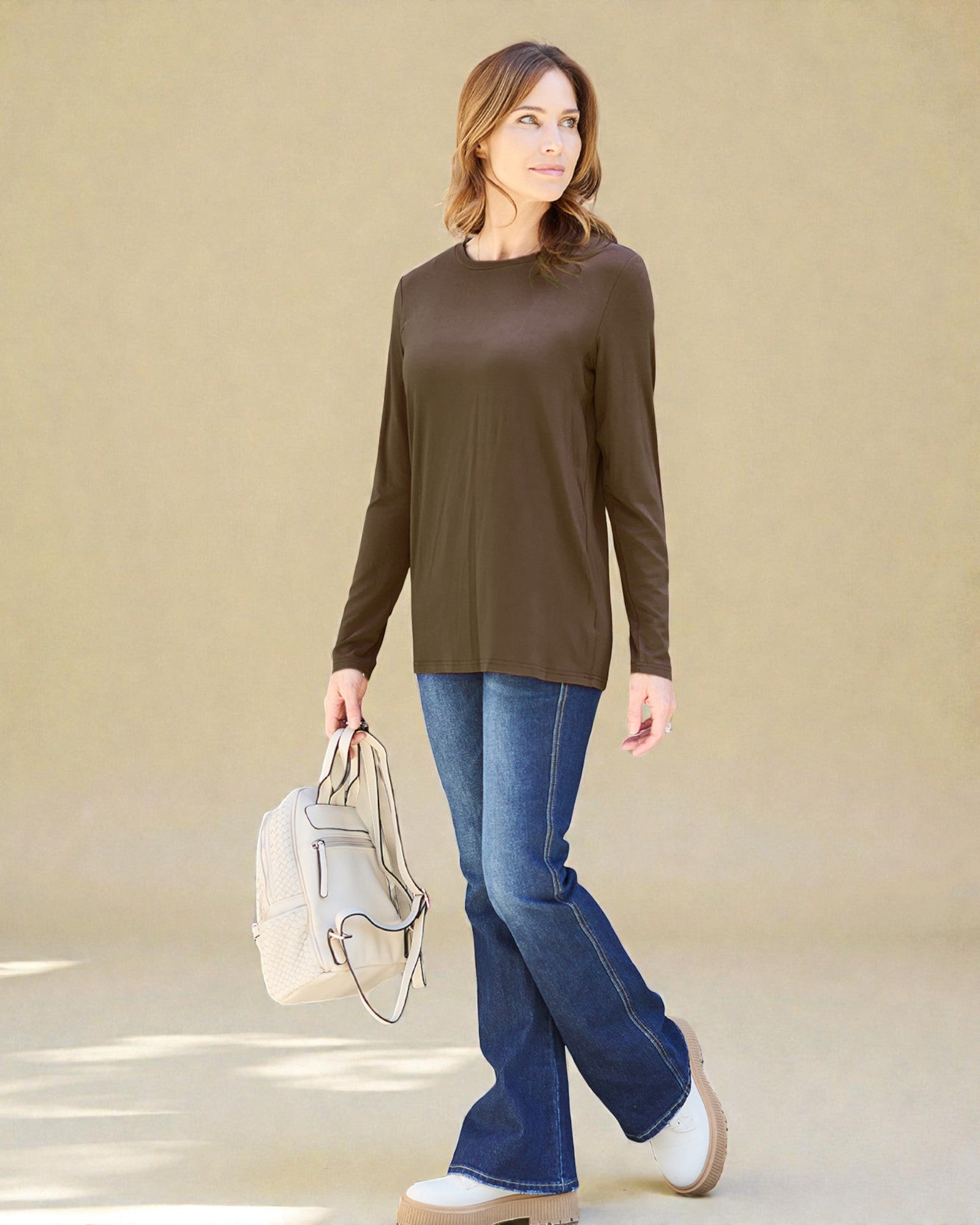 Woman wearing a brown long-sleeve top and blue jeans, holding a white backpack against a beige background
