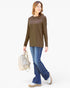 A woman modeling a long-sleeve crewneck top in olive brown, paired with blue jeans and white sneakers, while carrying a light beige shoulder bag against a plain white background.