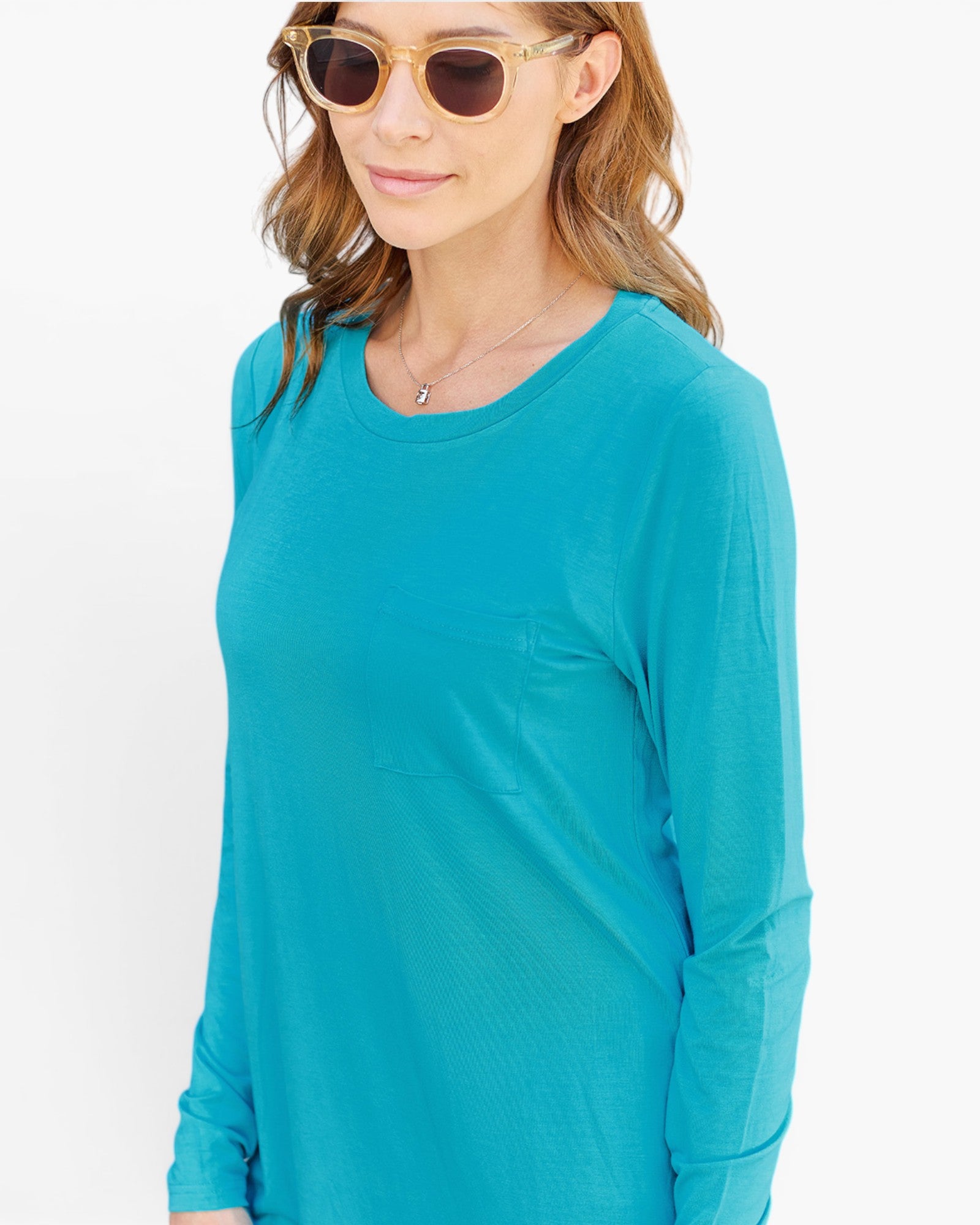 A woman wearing a bright turquoise long-sleeve crewneck top, gold-framed sunglasses, and a delicate necklace, shown against a plain white background.