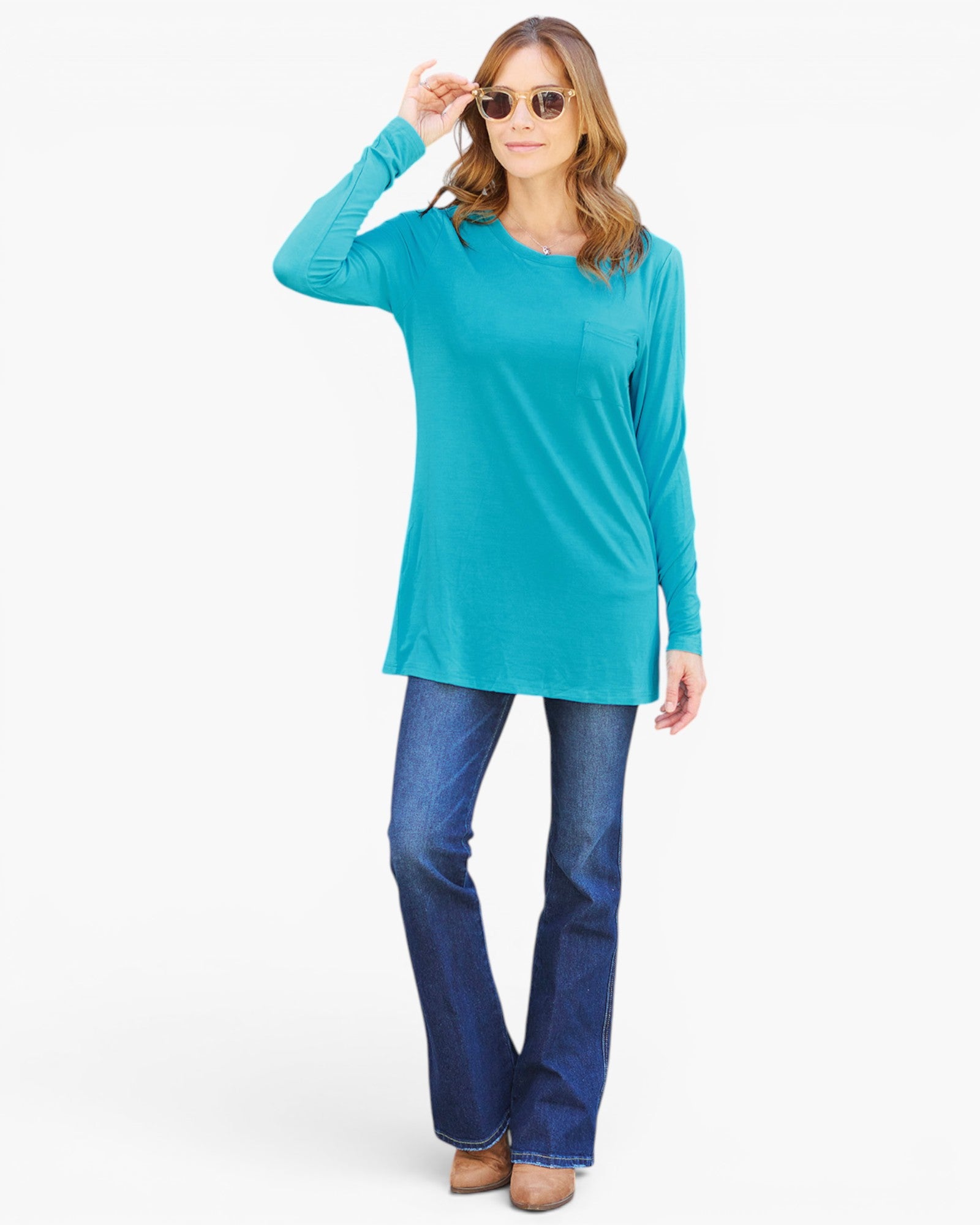A woman modeling a turquoise long-sleeve tunic top with blue jeans and sunglasses, standing against a plain white studio background.