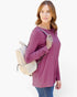 A woman wearing a dusty purple long-sleeve tunic top and blue jeans, carrying a cream woven backpack over her shoulder, set against a plain white background.