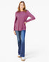 A woman modeling a long-sleeve dusty purple top with side ruching, paired with blue jeans and beige shoes, against a plain white studio background.