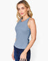 A woman modeling a sleeveless scoop-neck tank top in dusty blue, paired with dark blue jeans, against a plain white studio background.