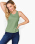 A woman modeling a sleeveless crew-neck tank top in sage green, paired with dark blue jeans, against a clean white background.