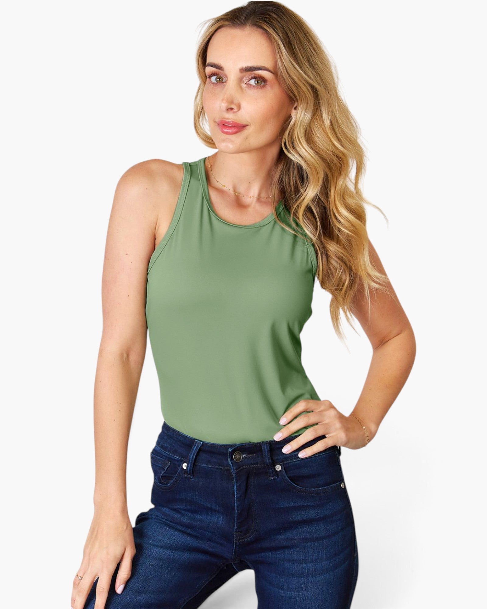 A woman modeling a sleeveless sage green tank top paired with dark blue jeans, photographed against a clean white studio background.