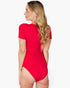 A woman with blonde wavy hair is shown from the back wearing a bright red short-sleeve one-piece swimsuit or bodysuit against a plain light gray studio background.