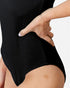 Close-up of a person wearing a black ribbed bodysuit or shapewear garment, with one hand resting on the hip against a light gray background.