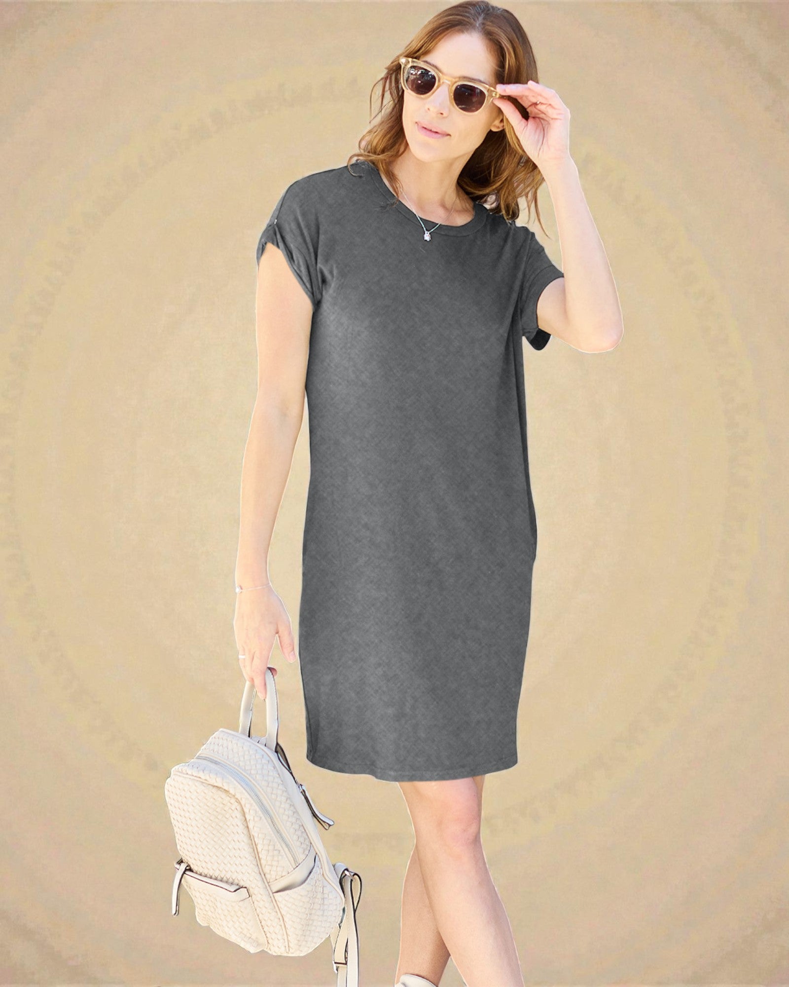 Woman wearing a gray t-shirt dress with sunglasses and a backpack on a beige background