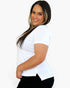 A woman with long dark hair and blonde highlights wearing a white short-sleeve t-shirt and black pants, posing against a solid white background.