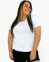 A woman with long dark hair wearing a plain white short-sleeve crewneck t-shirt and black shorts, smiling and looking to the side against a light gray background.