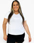 A woman modeling a plain white short-sleeve crewneck t-shirt paired with black shorts, set against a solid light gray background.