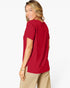 A woman wearing a short-sleeve red top and beige pants, shown from a side-back angle against a plain white studio background.