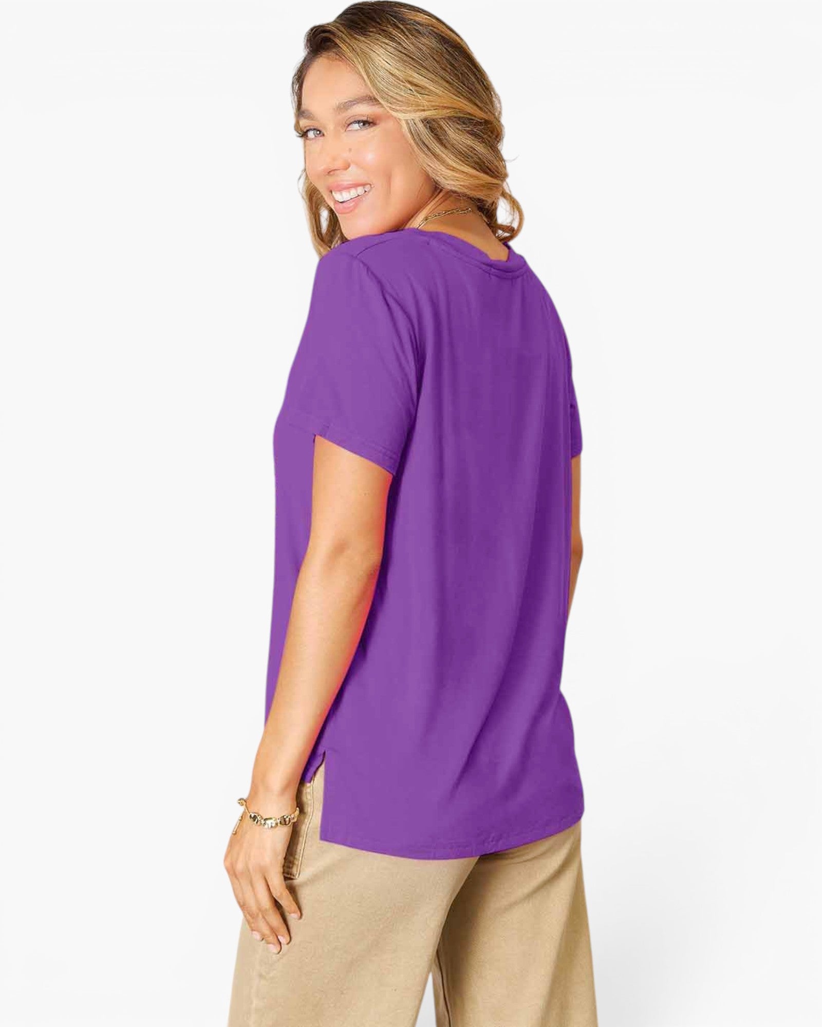 A woman wearing a purple short-sleeve t-shirt and beige pants, shown from a side-back angle against a clean white studio background.
