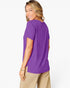 A woman wearing a purple short-sleeve t-shirt and beige pants, shown from a side-back angle against a clean white studio background.