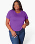 A woman modeling a short-sleeve crewneck t-shirt in solid purple, paired with dark blue jeans, against a light gray studio background.