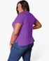 A woman wearing a purple short-sleeve top and blue jeans, photographed from the back against a light gray studio background.