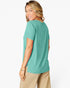 A woman wearing a short-sleeve teal t-shirt and beige pants, shown from a side-back angle against a plain white studio background.
