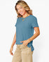 A woman wearing a short-sleeve blue t-shirt and beige pants, posing against a light gray studio background.