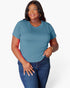 A woman modeling a short-sleeve crewneck t-shirt in a muted teal blue color, paired with dark blue jeans, against a plain light gray background.