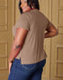 Person wearing a brown t-shirt and blue jeans standing against a beige wall.