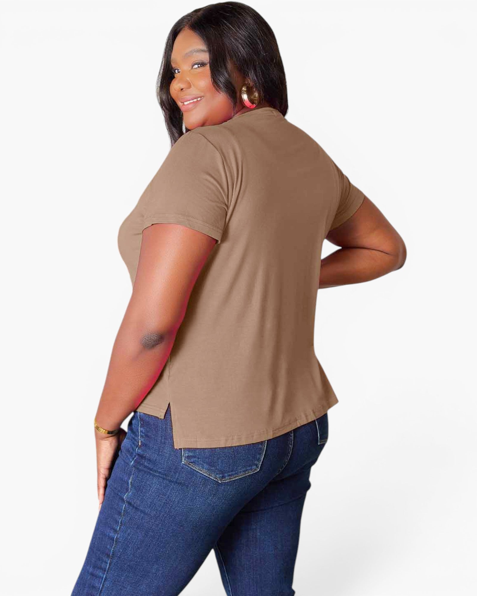A woman wearing a tan short-sleeve t-shirt and blue jeans, shown from a back-three-quarter angle against a light gray studio background.