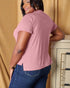 Woman wearing a pink t-shirt and blue jeans standing against a beige curtain.