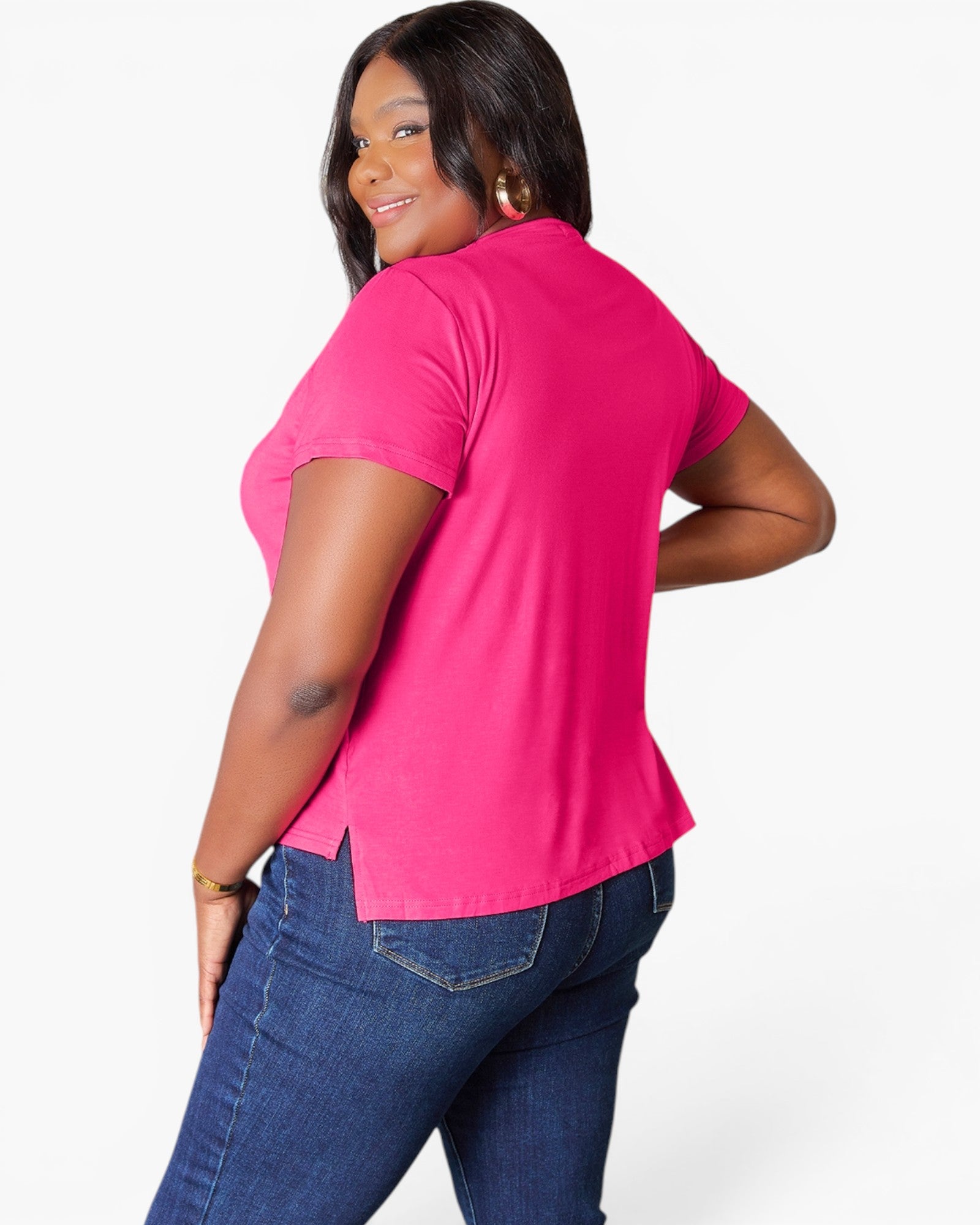 A woman wearing a bright pink short-sleeve top and dark blue jeans, shown from a side-back angle against a plain white studio background.