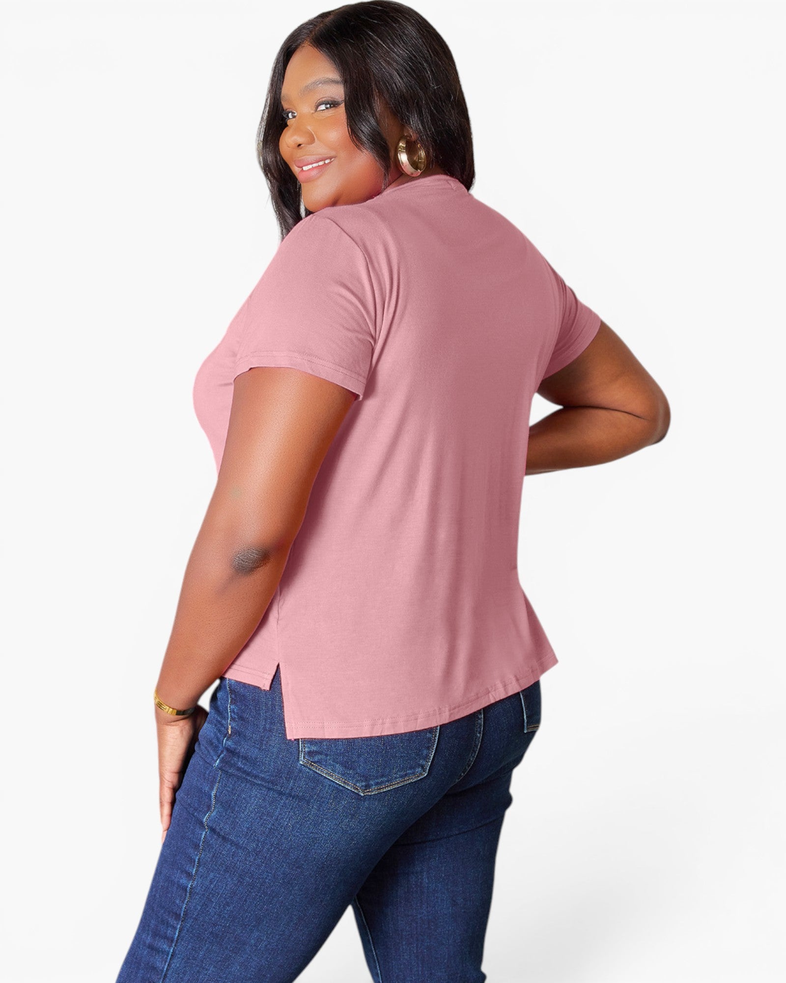 A woman wearing a dusty pink short-sleeve top and blue jeans, shown from a back-three-quarter angle against a light gray studio background.