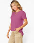 A woman wearing a short-sleeve purple top and beige pants, standing against a light gray studio background.