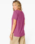 A woman wearing a short-sleeve crewneck t-shirt in a dusty magenta-purple color, paired with beige pants, photographed from a side-back angle against a clean white background.