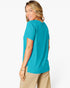 A woman wearing a bright turquoise short-sleeve t-shirt and beige pants, shown from a side-back angle against a clean white studio background.