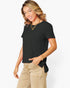 A woman modeling a black short-sleeve top with a side tie detail, paired with beige pants, against a plain white background.