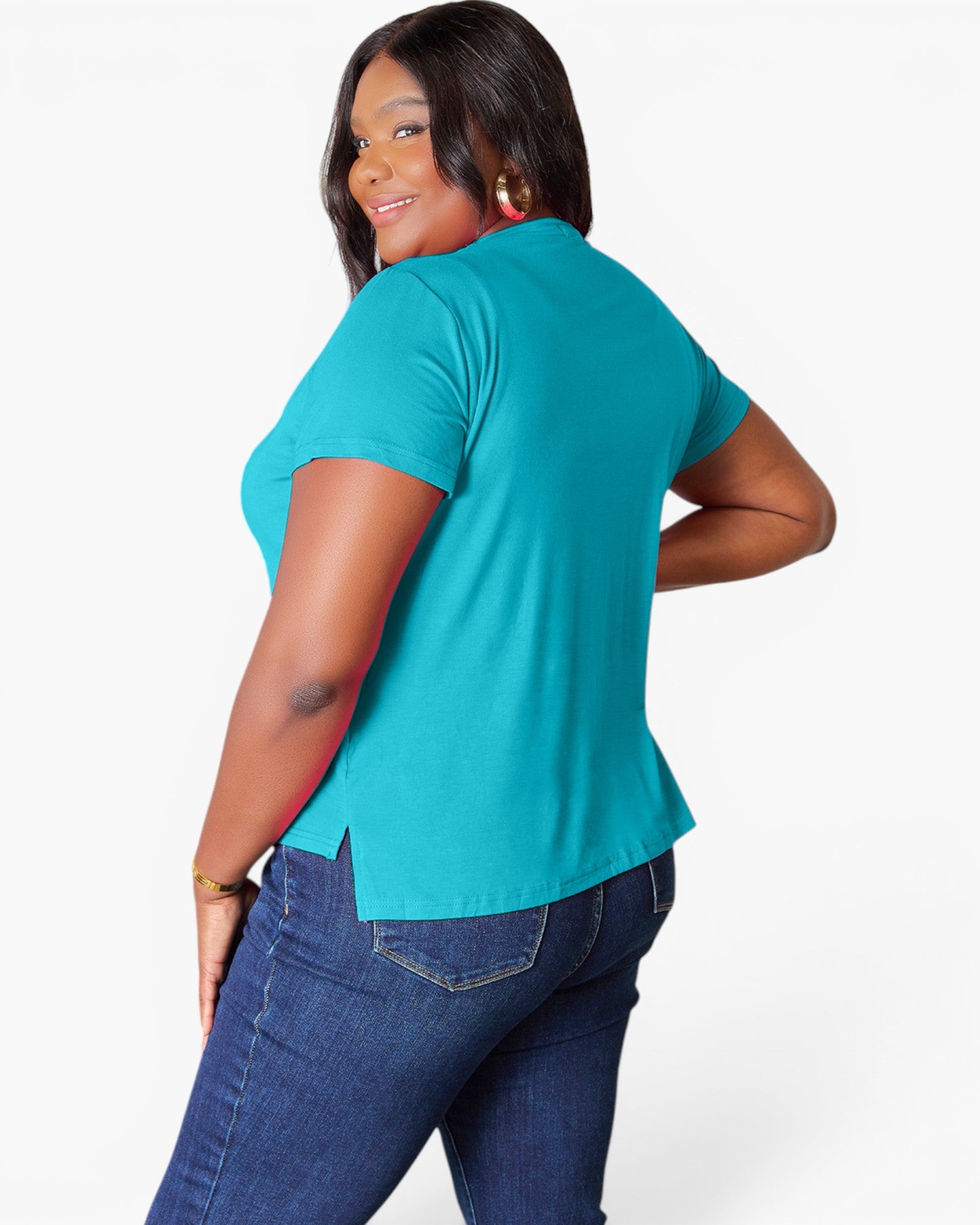 A woman wearing a bright turquoise short-sleeve top and dark blue jeans, photographed from the back against a clean white studio background.