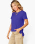 A woman modeling a short-sleeve royal blue top with a side tie detail, paired with beige pants, against a plain white studio background.