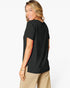 A woman wearing a black short-sleeve t-shirt and beige pants, photographed from the back-three-quarter angle against a clean white studio background.