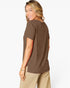 A woman wearing a brown short-sleeve t-shirt and beige pants, shown from a side-back angle against a plain white studio background.