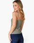A woman shown from the back wearing a sage green spaghetti-strap camisole top and dark blue jeans, set against a plain light gray studio background.