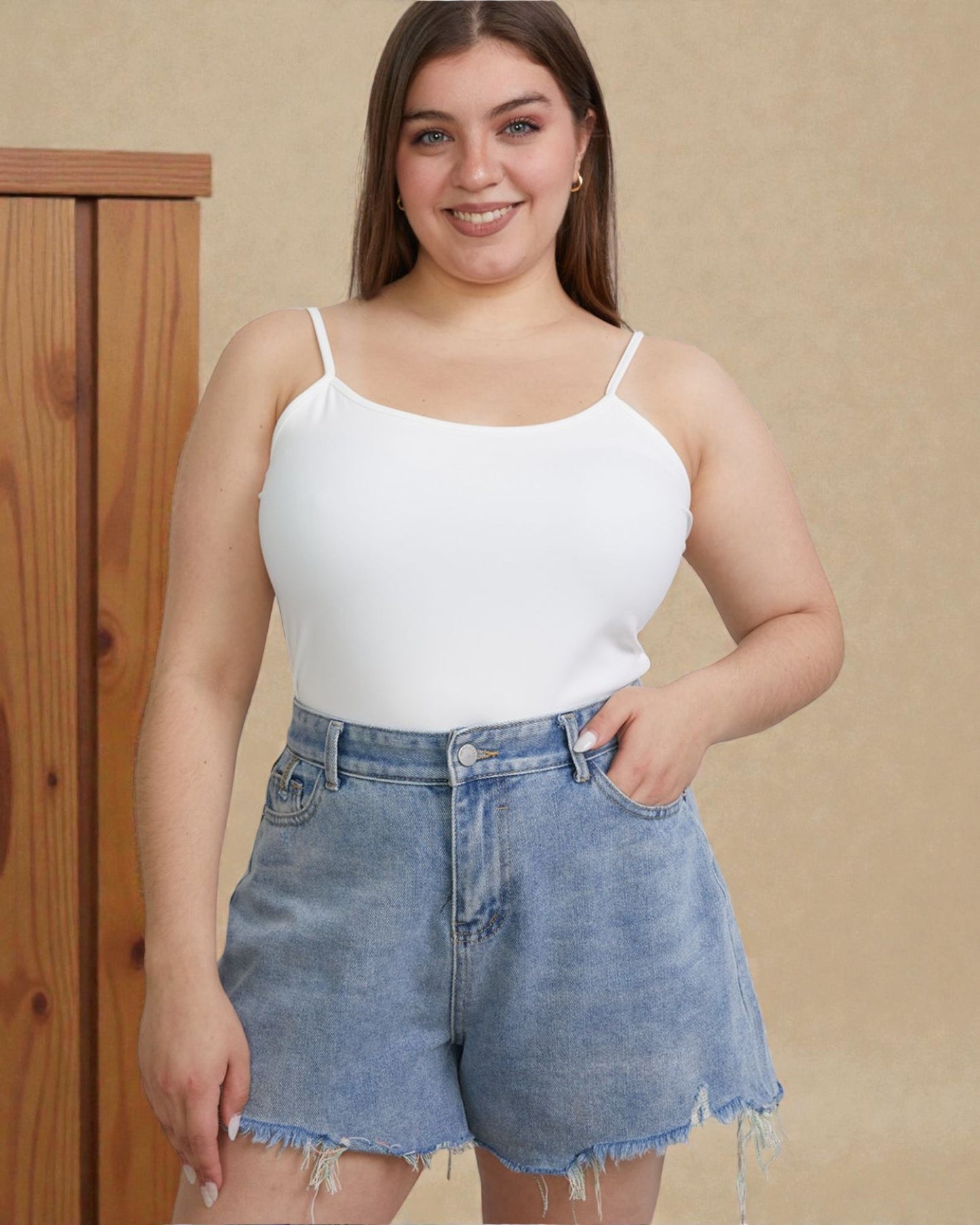 Woman wearing a white tank top and denim shorts against a beige wall.